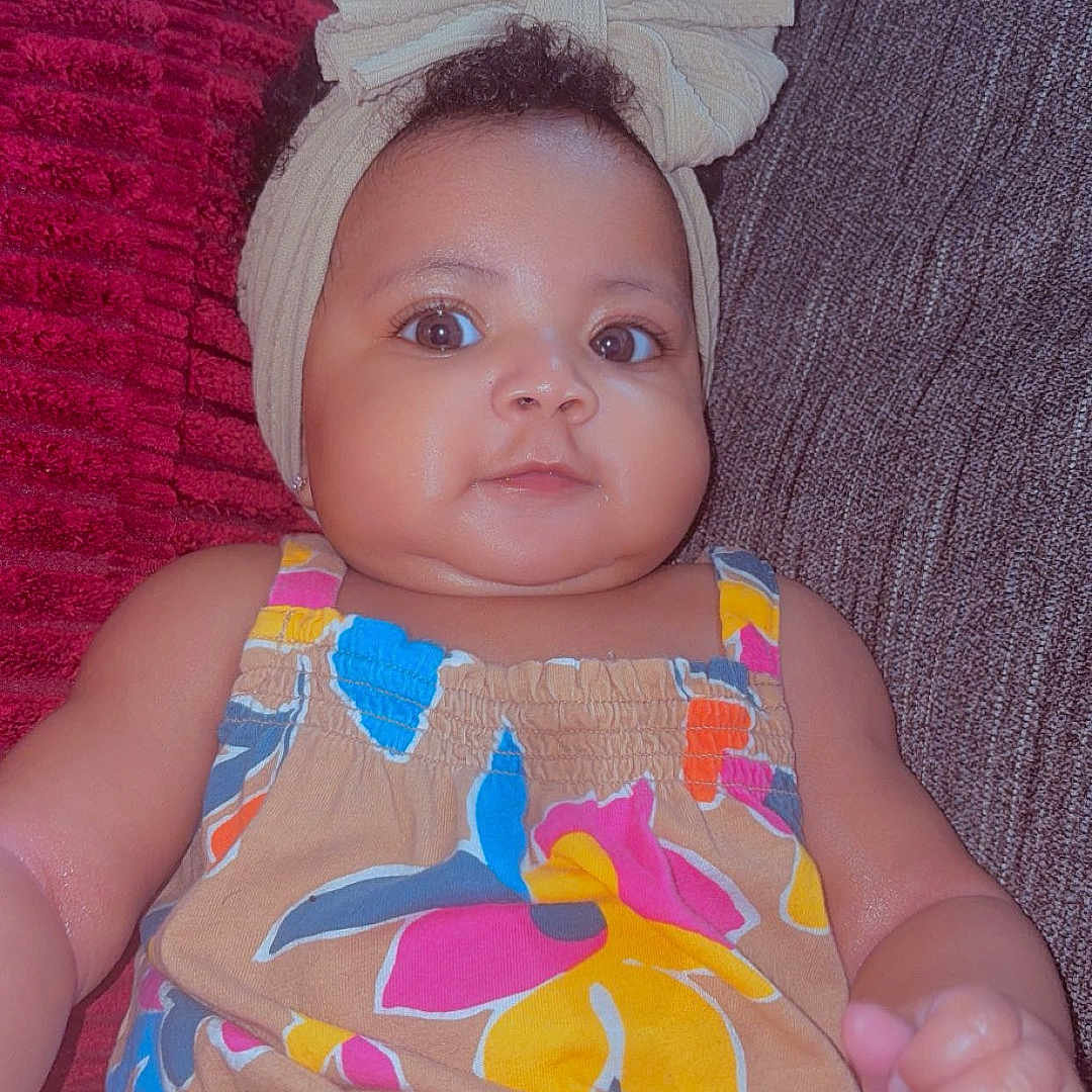 Naziya is registered to the contest to win money with this photo: baby, child, headband, bow, romper, colorful_clothing, couch, pillow, indoor, portrait, smiling, curious, infant, cute, person, skin, face, eyes, hand, seated