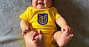 Alahia is registered to the contest to win money with this photo: baby, infant, smiling, yellow_onesie, feet, face, happy, lying_down, blanket, texture, child, cute, adorable, skin, head, person, small_feet, clothing, indoor, playful