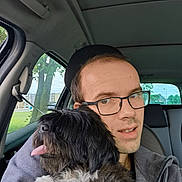 Hina participe au concours pour gagner de l'argent avec cette photo : man, glasses, dog, black_dog, white_fur, car_interior, seat, window, tree, buildings, tongue_out, pet, companion, casual_clothing, seatbelt, daylight, portrait, happy, closeup, indoors