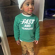 Khamari joined the competition — help win amazing prizes! child, toddler, indoor, beanie, green_sweatshirt, text_on_clothing, pants, white_sneakers, wooden_floor, living_room, couch, curious_expression, standing, clothing, person, young_child, casual_wear, floor, headwear, portrait