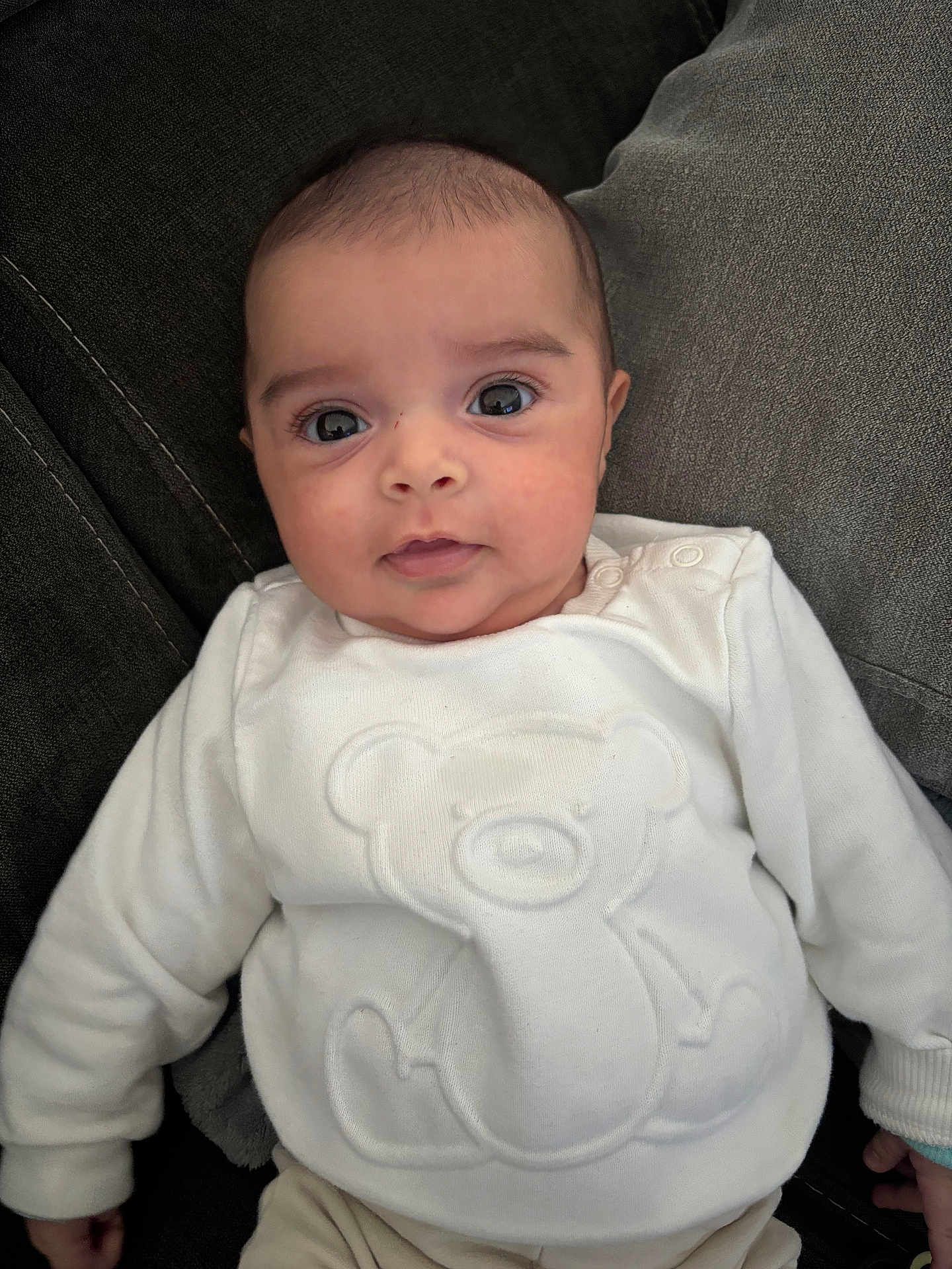 Aline participe au concours pour gagner de l'argent avec cette photo : baby, infant, white_sweater, teddy_bear_design, couch, cushion, soft_fabric, indoors, portrait, cute, child, person, smile, wide_eyes, young_child, sitting, cozy, comfortable, casual_clothing, relaxed