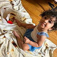 Egypt joined the competition — help win amazing prizes! child, toddler, baby, curly_hair, smiling, portrait, sitting, blanket, food, snack_box, indoors, wooden_floor, bed, furniture, dress, blue_clothing, barefoot, happy, close_up, home