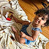 child, toddler, baby, curly_hair, smiling, portrait, sitting, blanket, food, snack_box, indoors, wooden_floor, bed, furniture, dress, blue_clothing, barefoot, happy, close_up, home
