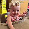 baby, child, high_chair, bow, headband, striped_clothing, hand, smile, face, indoor, furniture, caution_sign, wet_floor_sign, cabinet, tile_floor, cute, person, tongue_out, happy, playful