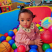 Lulu is registered to the contest to win money with this photo: baby, child, toddler, portrait, sitting, ball_pit, colorful_balls, basketball, toys, playroom, indoor, pink_dress, collar, curly_hair, wide_eyes, looking_at_camera, infant, play_area, colorful, happy