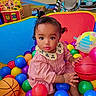 baby, child, toddler, portrait, sitting, ball_pit, colorful_balls, basketball, toys, playroom, indoor, pink_dress, collar, curly_hair, wide_eyes, looking_at_camera, infant, play_area, colorful, happy