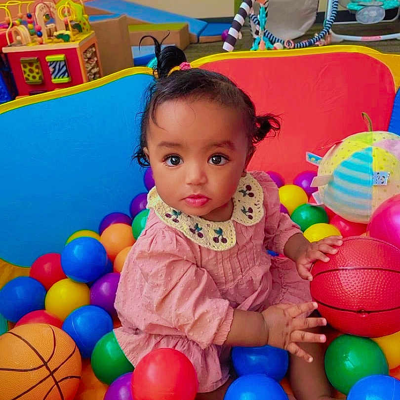 Lulu is registered to the contest to win money with this photo: baby, child, toddler, portrait, sitting, ball_pit, colorful_balls, basketball, toys, playroom, indoor, pink_dress, collar, curly_hair, wide_eyes, looking_at_camera, infant, play_area, colorful, happy