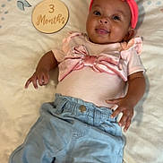 Càliyah is registered to the contest to win money with this photo: baby, infant, child, pink_headband, bow, blue_pants, blanket, floral_pattern, wooden_plaque, 3_months, smiling, cute, milestone, lying_down, clothing, happy, indoors, portrait, person, young_child