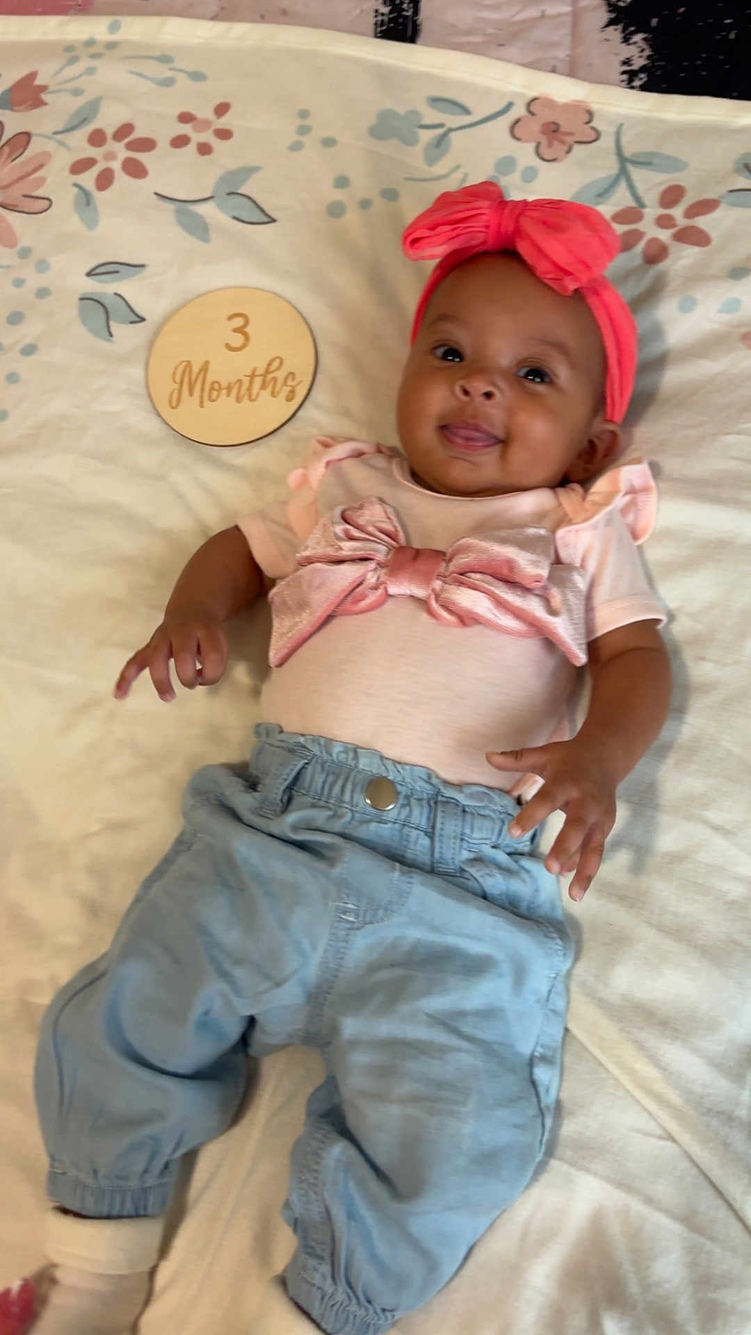 Càliyah is registered to the contest to win money with this photo: baby, infant, child, pink_headband, bow, blue_pants, blanket, floral_pattern, wooden_plaque, 3_months, smiling, cute, milestone, lying_down, clothing, happy, indoors, portrait, person, young_child