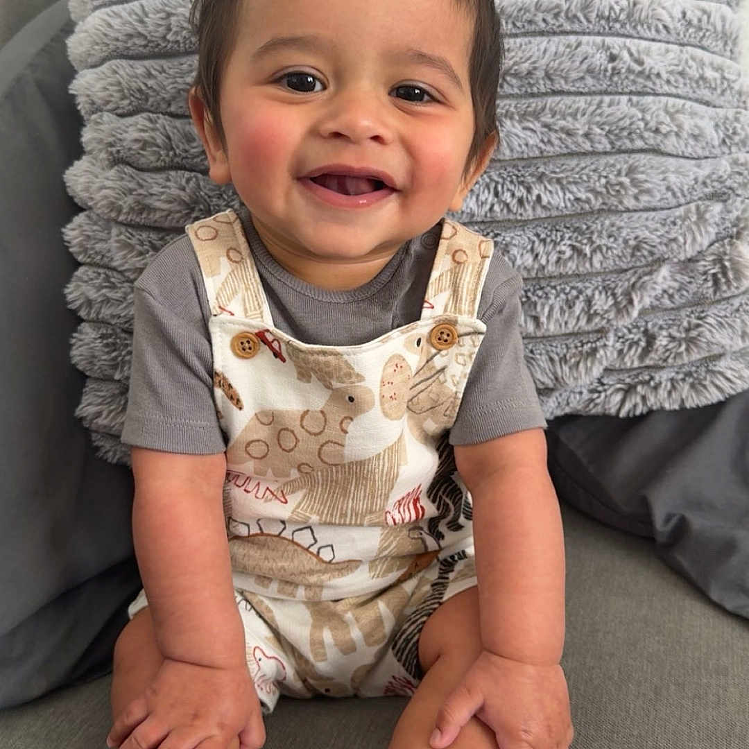 Zakkariya joined the competition — help win amazing prizes! baby, child, clothing, couch, cozy, cute, face, hands, happy, home, indoor, infant, legs, overalls, person, pillow, portrait, sitting, smiling, sock