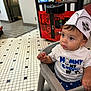 Julian is registered to the contest to win money with this photo: toddler, child, high_chair, paper_hat, steak_n_shake, tshirt, indoor, floor_tiles, claw_machine, plush_toys, restaurant, curious, seated, portrait, baby, clothing, person, young_child, casual, expression