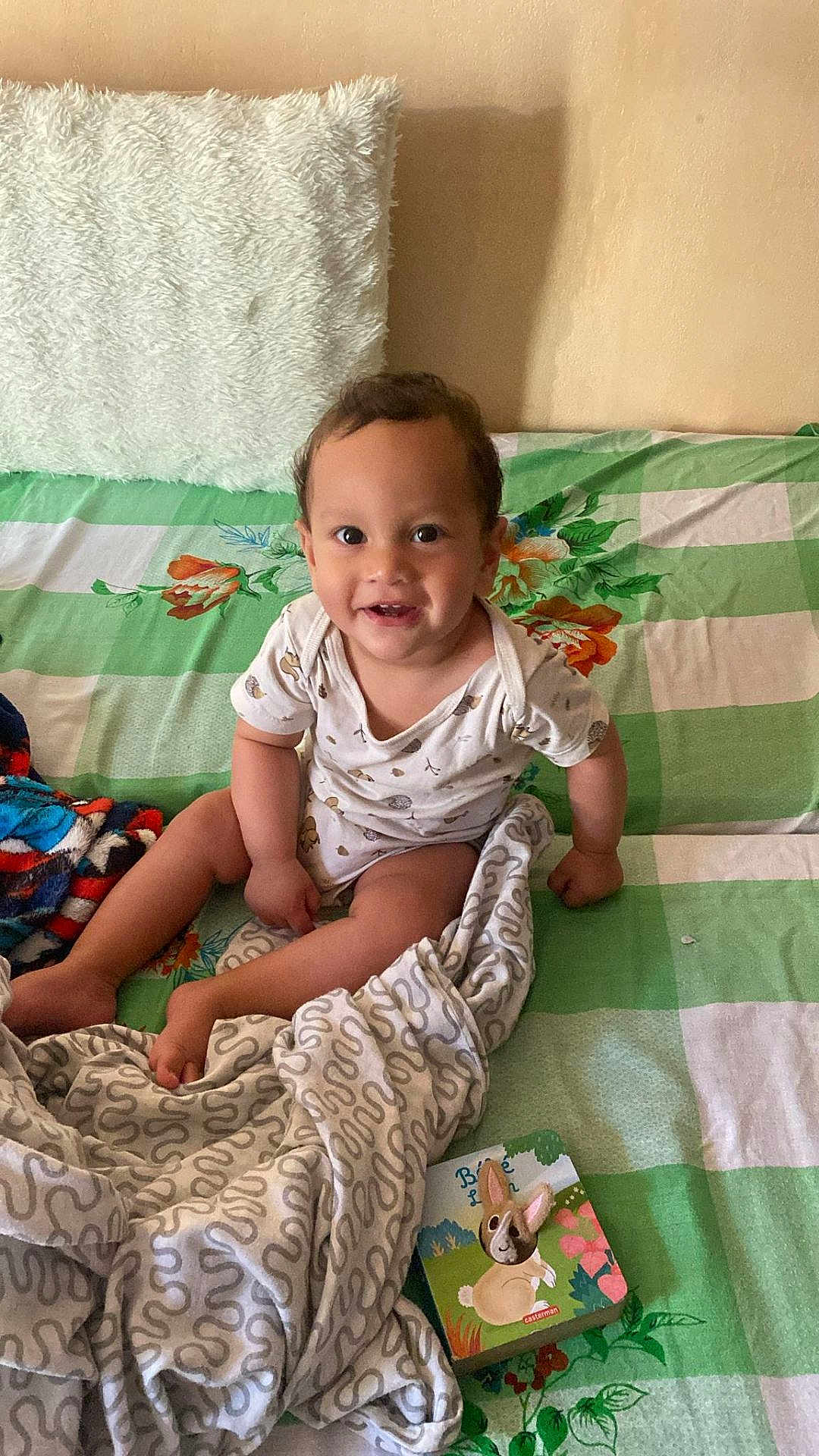 Kris Naidu Grandjean participe au concours pour gagner de l'argent avec cette photo : toddler, child, smiling, bed, blanket, book, toy, indoor, happy, person, face, clothing, fabric, pillow, floral_pattern, green, white, cozy, cute, portrait