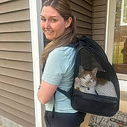 Taco is registered to the contest to win money with this photo: woman, smiling, cat, cat_in_backpack, backpack, pet_carrier, porch, home_exterior, vinyl_siding, door, window, shorts, tshirt, portrait, pet, happy, welcome_mat, rocks, pavement, daylight
