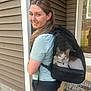 woman, smiling, cat, cat_in_backpack, backpack, pet_carrier, porch, home_exterior, vinyl_siding, door, window, shorts, tshirt, portrait, pet, happy, welcome_mat, rocks, pavement, daylight