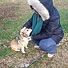 person, dog, park, grass, tree, jacket, hood, scarf, outdoor, petting, crouching, cold_weather, small_dog, nature, path, leafless_tree, casual_clothing, daytime, animal, human_animal_interaction