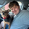 Bandit is registered to the contest to win money with this photo: dog, person, portrait, selfie, car, vehicle, indoor, seated, companion, pet, scruffy_dog, small_dog, red_collar, gray_shirt, brown_fur, face, smiling, road_trip, daylight, friendship