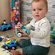 Sacha a rejoint le concours — aidez-le/la à gagner de superbes lots ! child, toddler, toy, play_mat, indoor, sitting, colorful_toys, building_blocks, car_toy, person, face, long_sleeve_shirt, pants, barefoot, floor, expression, wall, plastic_toys, cubby_storage, curious