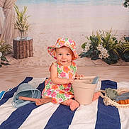 Layla is registered to the contest to win money with this photo: baby, child, floral_dress, sunhat, striped_towel, beach_toys, indoor, smiling, playing, bucket, shovel, flowers, plants, background, portrait, sitting, cute, happy, summer, toddler