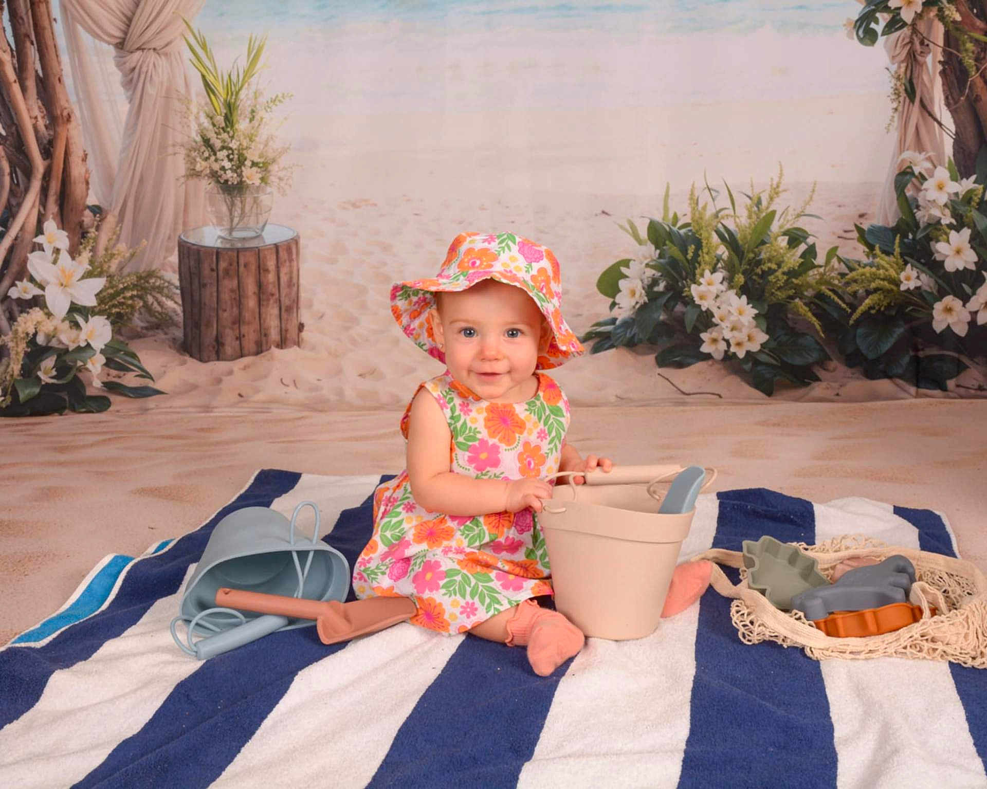 Layla is registered to the contest to win money with this photo: baby, child, floral_dress, sunhat, striped_towel, beach_toys, indoor, smiling, playing, bucket, shovel, flowers, plants, background, portrait, sitting, cute, happy, summer, toddler