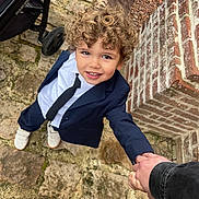 Erwan participe au concours pour gagner de l'argent avec cette photo : child, toddler, curly_hair, smiling, suit, tie, hand_holding, brick_wall, stone_path, outdoor, footwear, stroller, person, casual, happy, young_child, fashion, portrait, candid, daylight