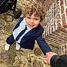brick_wall, candid, casual, child, curly_hair, daylight, fashion, footwear, hand_holding, happy, outdoor, person, portrait, smiling, stone_path, stroller, suit, tie, toddler, young_child