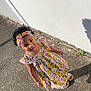 child, girl, dress, headband, floral_pattern, sidewalk, shadow, outdoor, sunlight, curly_hair, colorful, standing, concrete, wall, footwear, casual, expression, person, summer, daytime