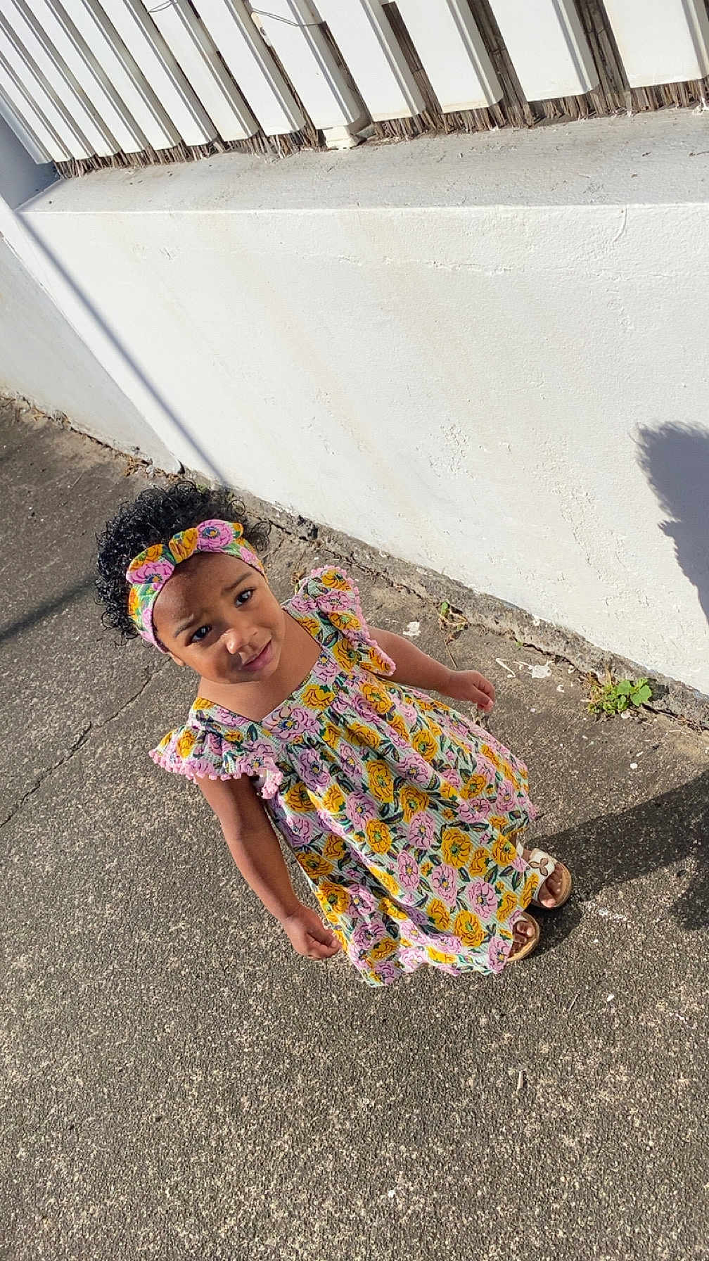 Aëlyah a rejoint le concours — aidez-le/la à gagner de superbes lots ! child, girl, dress, headband, floral_pattern, sidewalk, shadow, outdoor, sunlight, curly_hair, colorful, standing, concrete, wall, footwear, casual, expression, person, summer, daytime