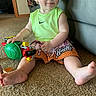 Zane is registered to the contest to win money with this photo: toddler, child, toy, carpet, indoor, clothing, shorts, sleeveless_shirt, barefoot, smiling, face, person, playful, sitting, home, floor, colorful, green, red, cute