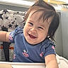 Lily Emelia joined the competition — help win amazing prizes! baby, child, smile, high_chair, blue_shirt, pony_tail, indoor, window_blinds, sunlight, happy, cute, infant, arm, hand, face, person, young_child, clothing, seat, background