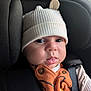 baby, infant, hat, car_seat, black, orange_clothing, buttons, face, eyes, mouth, tongue, ribbed_hat, cute, child, seat_belt, closeup, indoors, young_child, person, apparel