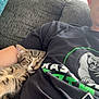 Bug is registered to the contest to win money with this photo: cat, tabby_cat, sleeping, person, black_tshirt, graphic_tshirt, couch, gray_couch, arm, indoor, relaxing, pet, cute, cozy, nap, resting, feline, domestic_animal, companion, home