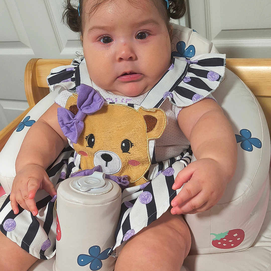 Carola is registered to the contest to win money with this photo: baby, baby_feet, chair, child, colorful, cup, dress, face, hair_bows, high_chair, home, indoor, infant, portrait, purple_bows, sitting, smiling, striped_clothing, teddy_bear, toy