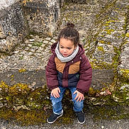 Maël participe au concours pour gagner de l'argent avec cette photo : archway, casual_clothing, child, cobblestone, curly_hair, gray_scarf, historic_site, jeans, moss, nature, outdoor, puffer_jacket, seated, sneakers, stone_path, stone_wall, thoughtful, urban_decay, weathered_stone, young_person