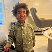 Maël a rejoint le concours — aidez-le/la à gagner de superbes lots ! child, smiling, indoor, curly_hair, green_jacket, living_room, rocking_chair, coffee_table, floor, person, portrait, happy, furniture, casual_clothing, young_child, home, standing, face, cute, bright