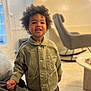 bright, casual_clothing, child, coffee_table, curly_hair, cute, face, floor, furniture, green_jacket, happy, home, indoor, living_room, person, portrait, rocking_chair, smiling, standing, young_child