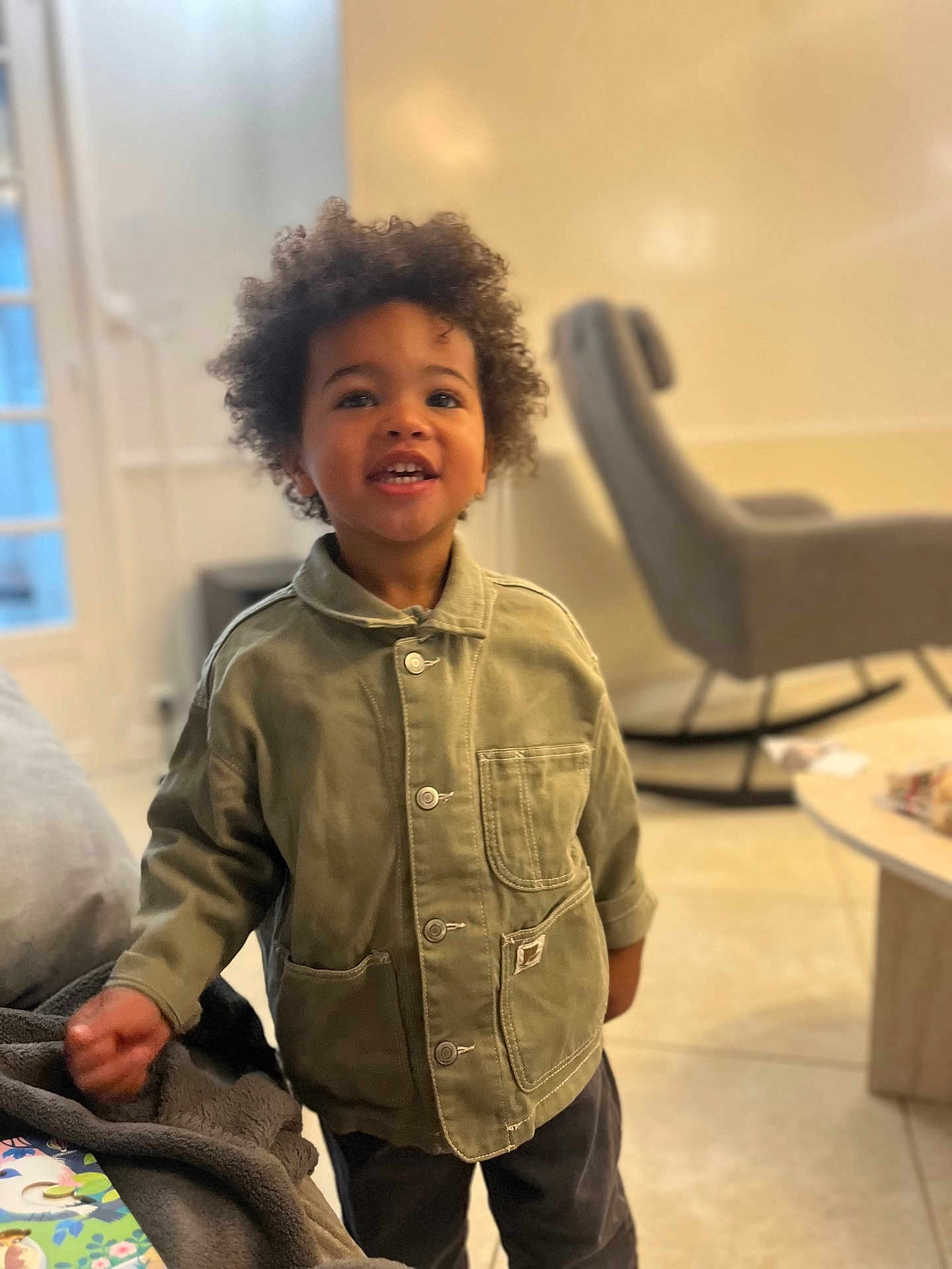 Maël a rejoint le concours — aidez-le/la à gagner de superbes lots ! child, smiling, indoor, curly_hair, green_jacket, living_room, rocking_chair, coffee_table, floor, person, portrait, happy, furniture, casual_clothing, young_child, home, standing, face, cute, bright
