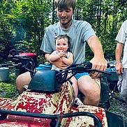 Emma Jean is registered to the contest to win money with this photo: baby, child, man, atv, outdoor, forest, greenery, summer, smile, clapping, shorts, gray_shirt, vehicle, nature, person, happy, trees, pigtails, grass, sunlight