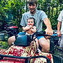 baby, child, man, atv, outdoor, forest, greenery, summer, smile, clapping, shorts, gray_shirt, vehicle, nature, person, happy, trees, pigtails, grass, sunlight