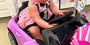 Emma Jean is registered to the contest to win money with this photo: toddler, child, toy_car, purple_car, pink_bows, dress, steering_wheel, reflection, woman, boots, white_boots, concrete_floor, indoor, play, cute, small_car, pink_bow, person, hair, legs