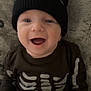 baby, smiling, beanie, black_clothing, skeleton_outfit, infant, happy, cute, face, headwear, child, lying_down, blanket, soft_texture, portrait, closeup, indoor, young, person, apparel
