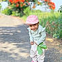 child, pink_cap, smiling, green_leaves, road, outdoor, nature, plants, orange_flowers, sandals, cute, playful, young_child, casual_clothing, sunlight, portrait, happy, trees, background_blur, standing