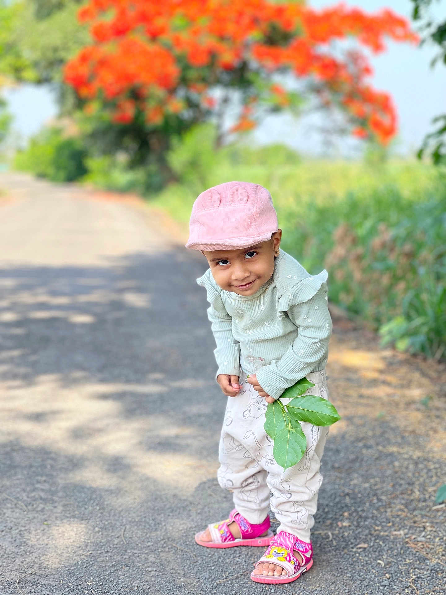 Ananya is registered to the contest to win money with this photo: child, pink_cap, smiling, green_leaves, road, outdoor, nature, plants, orange_flowers, sandals, cute, playful, young_child, casual_clothing, sunlight, portrait, happy, trees, background_blur, standing