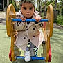 child, toddler, trench_coat, headwear, playground, spring_rider, outdoor, park, trees, daylight, shoes, blue_bar, yellow_wood, green_ground, curious, person, playing, young_child, casual_clothing, shadow
