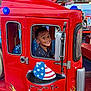 child, smiling, fire_truck, red_vehicle, amusement_ride, mickey_mouse, carnival, lights, toy_vehicle, happy, boy, face, window, seat, indoor, entertainment, fun, colorful, amusement_park, fire_rescue
