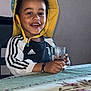 child, smiling, hat, yellow_hat, jacket, adidas, glass, table, tablecloth, floral_pattern, indoor, happy, sitting, person, curly_hair, black_hair, bracelet, youth, portrait, casual