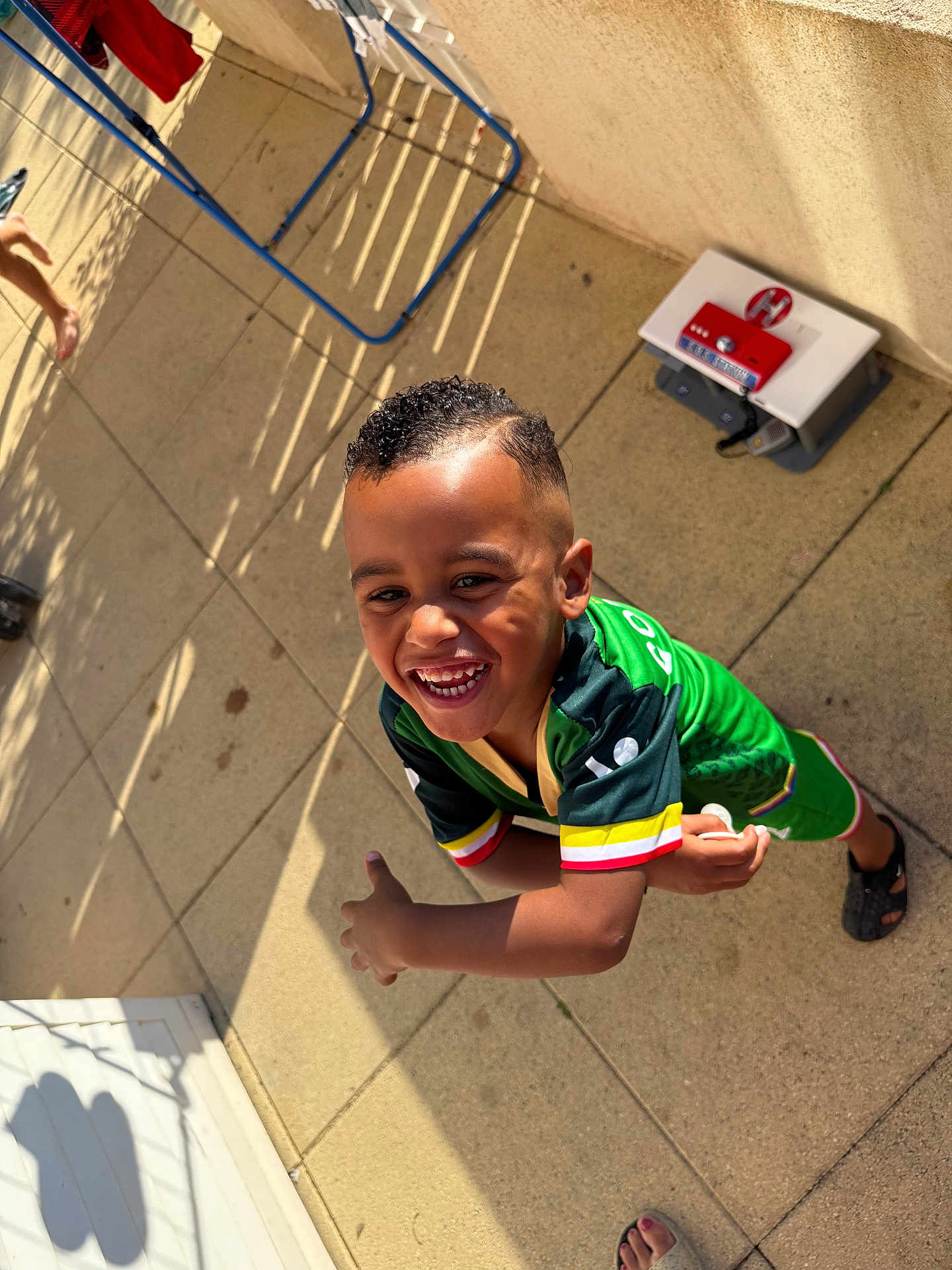 Nidhoimi Farrel a rejoint le concours — aidez-le/la à gagner de superbes lots ! child, smiling, green_clothing, short_hair, outdoor, sunlight, shadow, tile_floor, barefoot, sportswear, happy, boy, playful, patio, casual, daylight, person, expression, motion, background