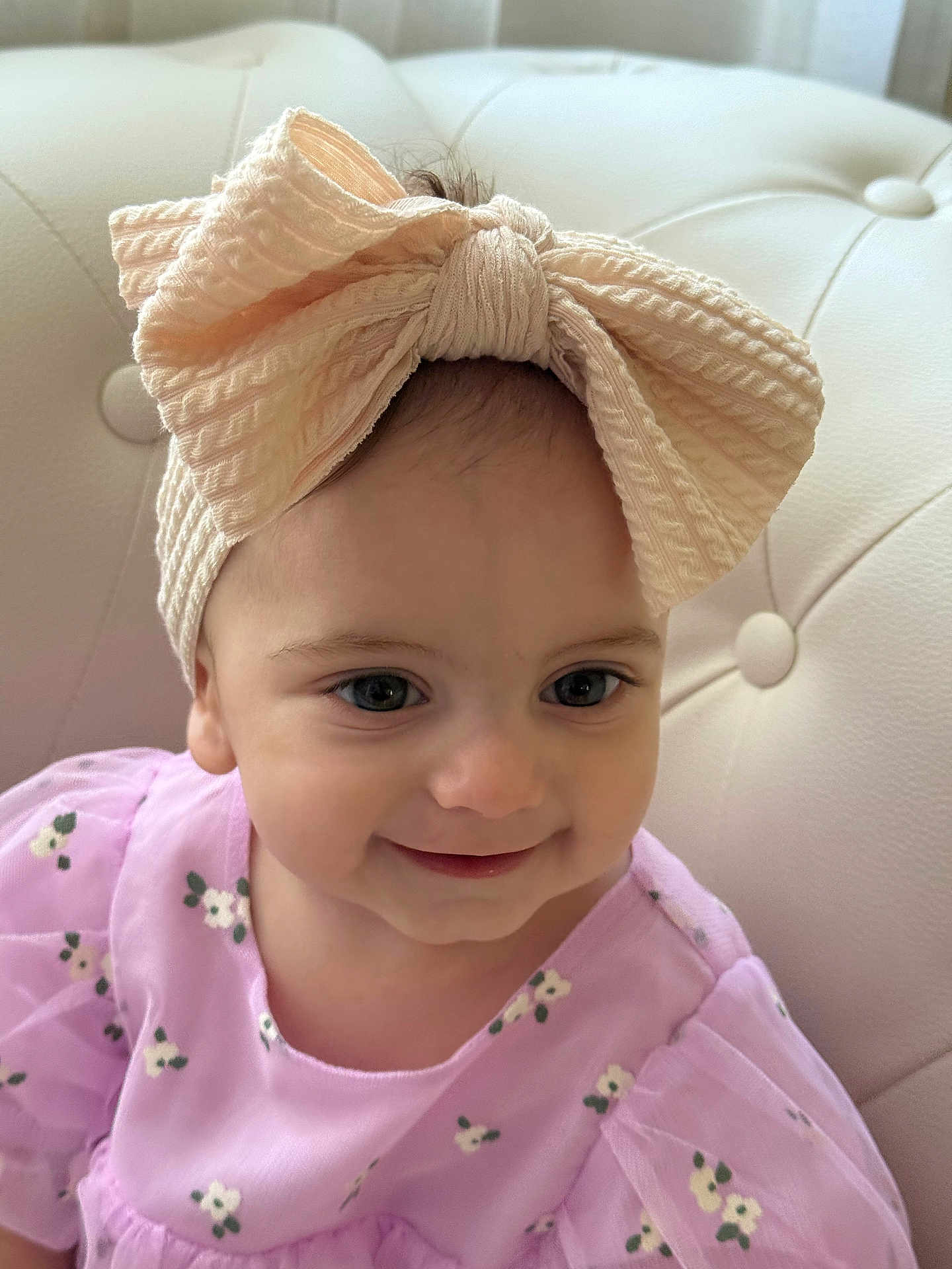 Aida is registered to the contest to win money with this photo: baby, child, headband, bow, smile, face, eyes, dress, floral, purple, couch, upholstery, toddler, portrait, cute, indoors, soft_light, happy, person, fashion