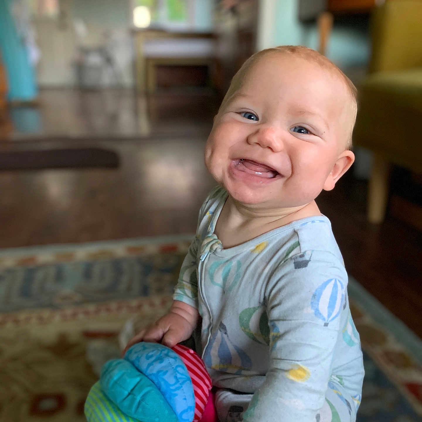 Hunter is registered to the contest to win money with this photo: baby, blue_eyes, cheerful, child, colorful, cute, face, floor, happy, home, indoor, infant, pajamas, patterned_rug, person, rug, sitting, smiling, soft_toy, toy