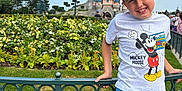 Noë a rejoint le concours — aidez-le/la à gagner de superbes lots ! child, boy, mickey_mouse, hat, tshirt, shorts, smile, flowers, flower_bed, castle, theme_park, fence, greenery, outdoor, daytime, sky, people, tourists, grass, happy