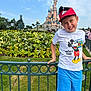 child, boy, mickey_mouse, hat, tshirt, shorts, smile, flowers, flower_bed, castle, theme_park, fence, greenery, outdoor, daytime, sky, people, tourists, grass, happy